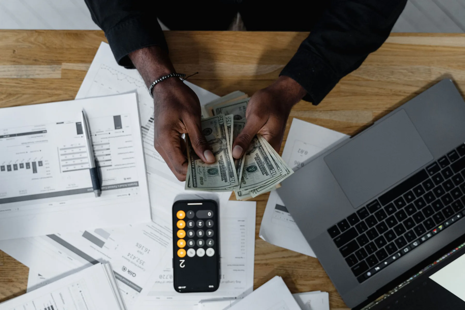 Hands counting dollars beside a laptop and calculator.